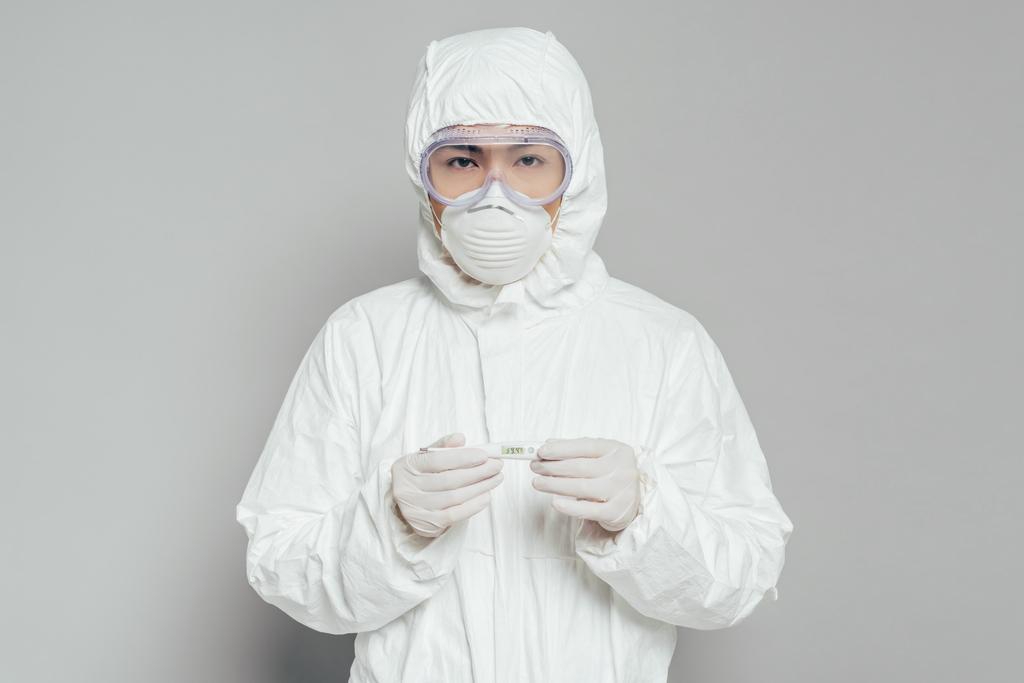 epidemiólogo asiático mirando cámara mientras sostiene termómetro mostrando alta temperatura sobre fondo gris
 - Foto, imagen