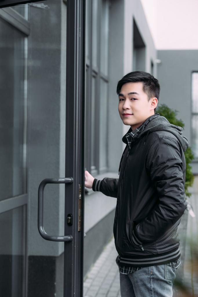 joven asiático hombre mirando cámara mientras de pie cerca de construcción y apertura de la puerta
 - Foto, Imagen