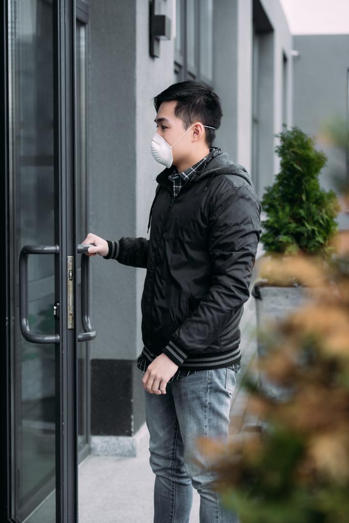valikoiva painopiste nuori aasialainen mies hengityssuojain naamio avaamalla ovi ja astumalla rakennukseen
 - Valokuva, kuva