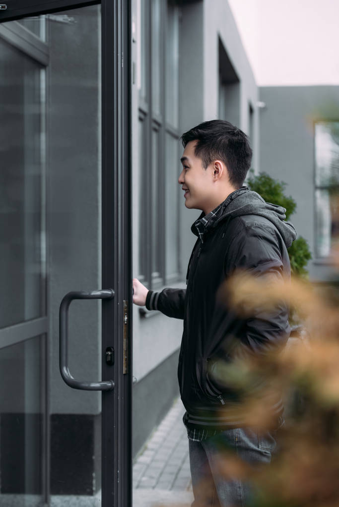 valikoiva painopiste nuori aasialainen mies hymyilee avatessaan oven ja astuessaan rakennukseen
 - Valokuva, kuva