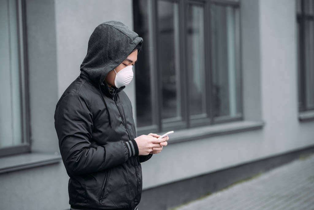 fiatal ázsiai férfi légzőkészülék maszk chat okostelefon, miközben áll az utcán - Fotó, kép