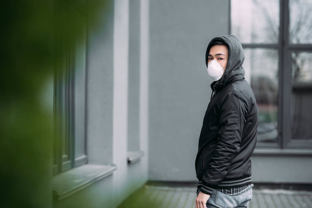 Selektiver Fokus eines jungen asiatischen Mannes mit Atemschutzmaske, der auf der Straße in die Kamera blickt - Foto, Bild