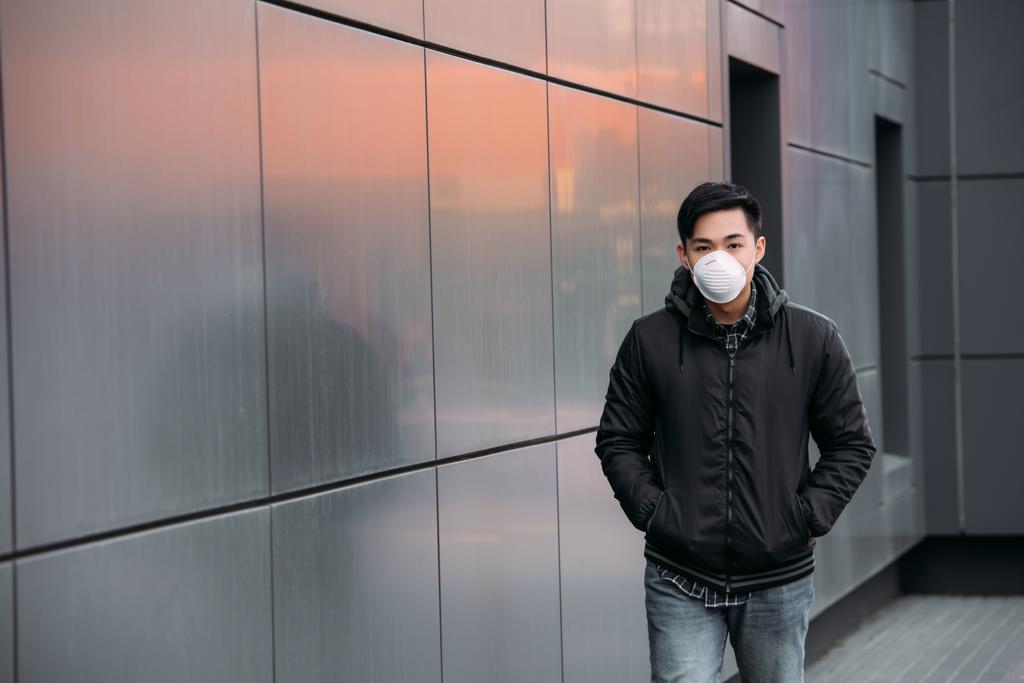 jonge aziatische man in respirator masker kijken naar camera tijdens het lopen langs de muur met de handen in zakken - Foto, afbeelding
