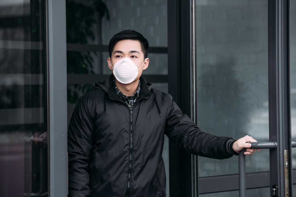 young asian man in respirator mask looking away while leaving building - 写真・画像