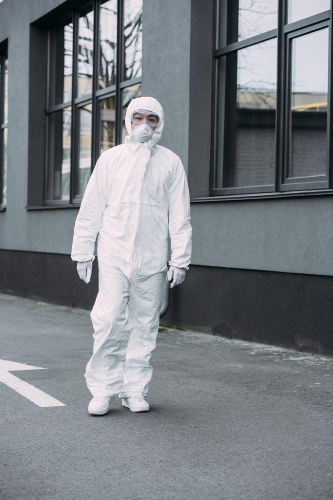 asian epidemiologist in hazmat suit and respirator mask looking at camera while standing on street near building - 写真・画像