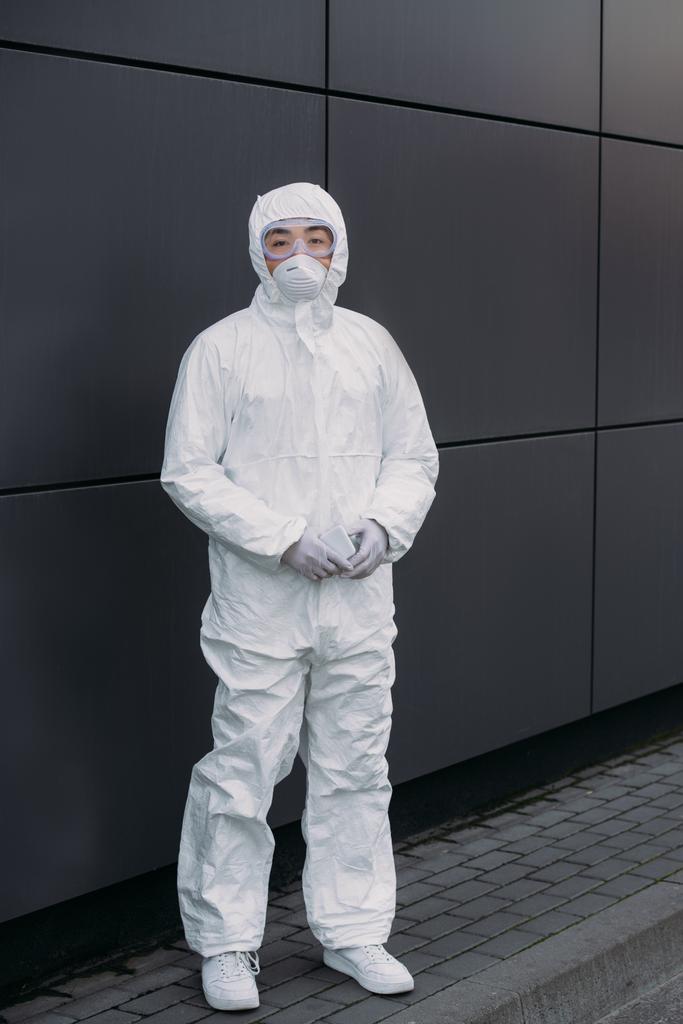 Asiatischer Epidemiologe in Warnanzug und Atemschutzmaske blickt in die Kamera, während er an der Wand steht - Foto, Bild