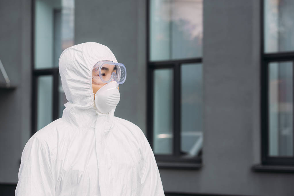 asian epidemiologist in hazmat suit and respirator mask looking away while standing near building - Fotoğraf, Görsel