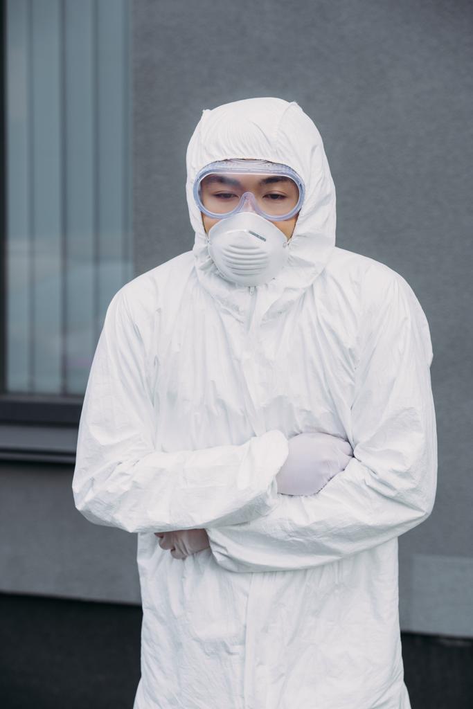 epidemiólogo asiático en traje de hazmat y máscara respiratoria de pie con los brazos cruzados mientras sufre de dolor abdominal sintomático en la calle
 - Foto, Imagen