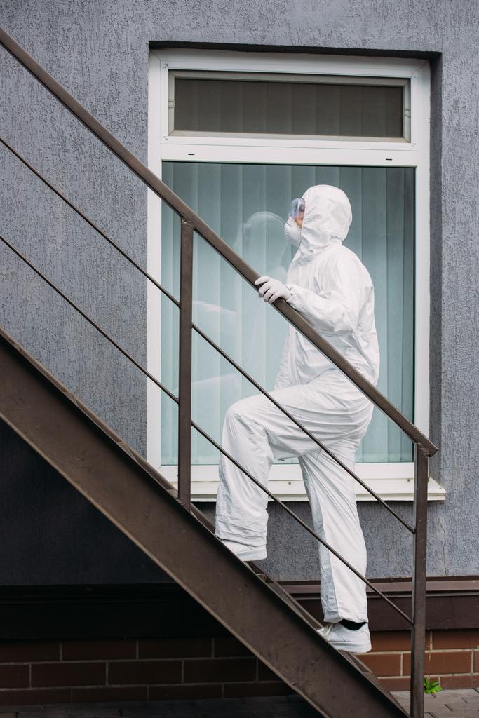 Эпидемиолог из Азии в защитном костюме и респираторной маске поднимается по лестнице к зданию
 - Фото, изображение
