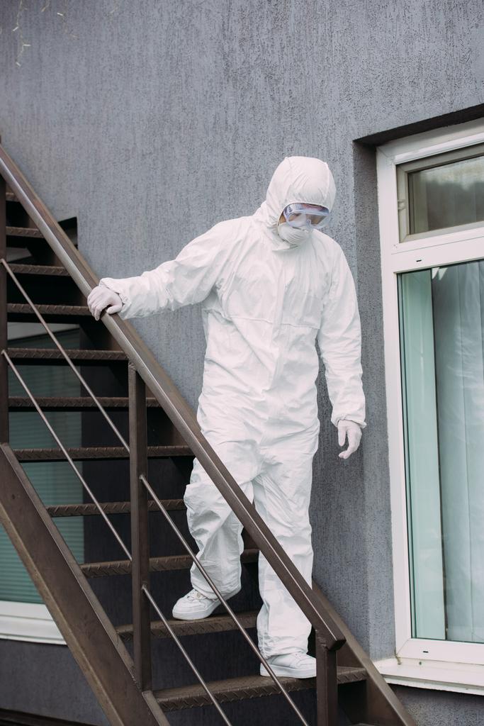 Азиатский эпидемиолог в защитном костюме и респираторной маске спускается вниз по лестнице здания
 - Фото, изображение