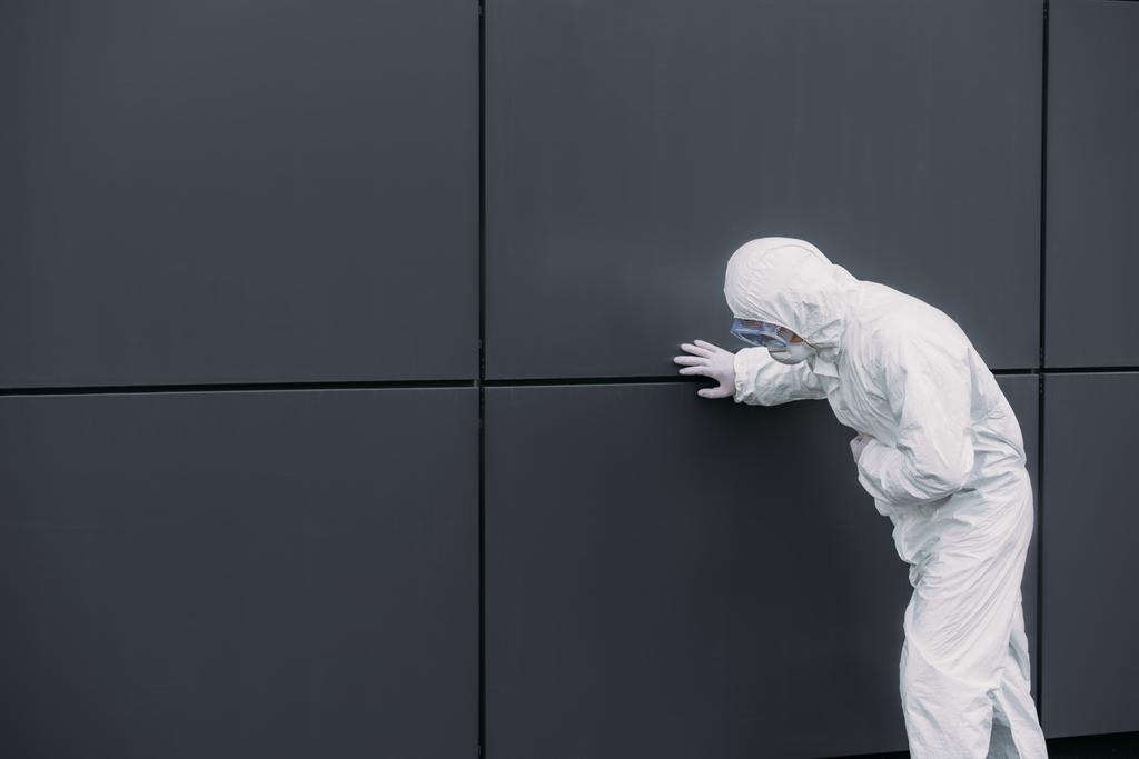 Азиатский эпидемиолог в защитном костюме опирается на стену, страдая от симптоматической боли в животе
 - Фото, изображение