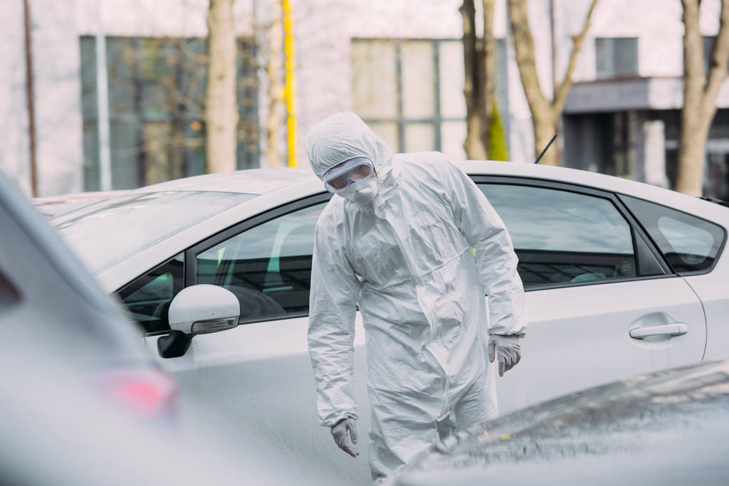 enfoque selectivo de epidemiólogo asiático en traje de materiales peligrosos y máscara respiratoria inspeccionar vehículos en el estacionamiento
 - Foto, imagen