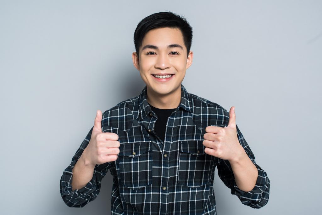 glimlachende aziatische man kijken naar camera terwijl het tonen van duimen tot op grijze achtergrond - Foto, afbeelding