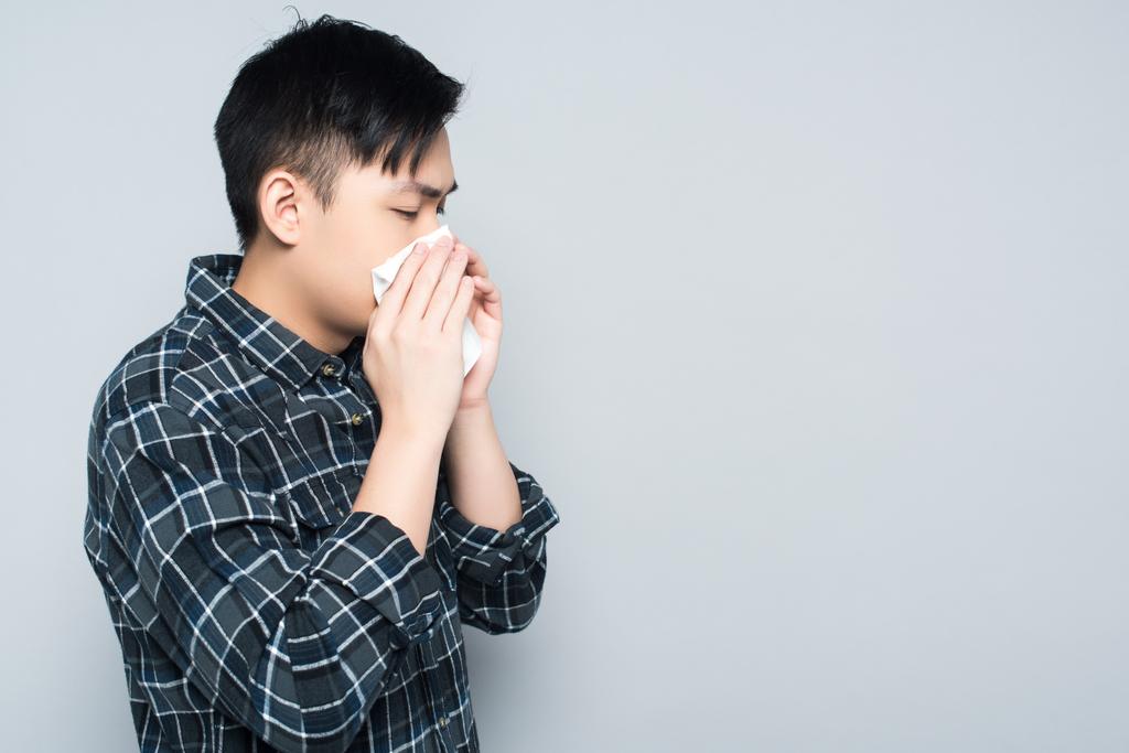 νεαρός Ασιάτης σκουπίζει τη μύτη του με χαρτοπετσέτα ενώ φτερνίζεται απομονωμένος σε γκρι - Φωτογραφία, εικόνα