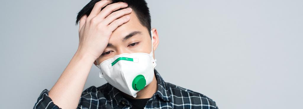 panorâmica tiro de jovem asiático homem no respirador máscara tocando testa enquanto sofrendo de dor de cabeça isolado no cinza
 - Foto, Imagem