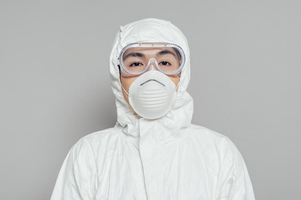 Asiatischer Epidemiologe in Warnanzug und Atemschutzmaske blickt in die Kamera auf grauem Hintergrund - Foto, Bild