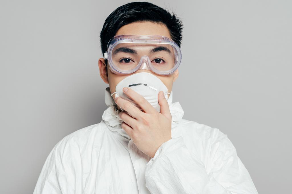 азиатский эпидемиолог в защитном костюме трогает респираторную маску, глядя на камеру, изолированную на сером
 - Фото, изображение