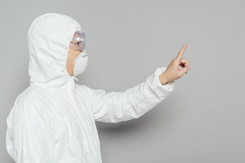 vista lateral del epidemiólogo asiático en traje de hazmat y máscara respiratoria mostrando gesto de advertencia sobre fondo gris
 - Foto, Imagen