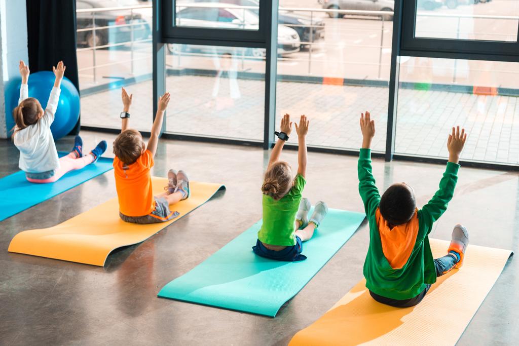 Visão traseira de crianças com mãos no ar exercitando-se em tapetes de fitness no ginásio
 - Foto, Imagem