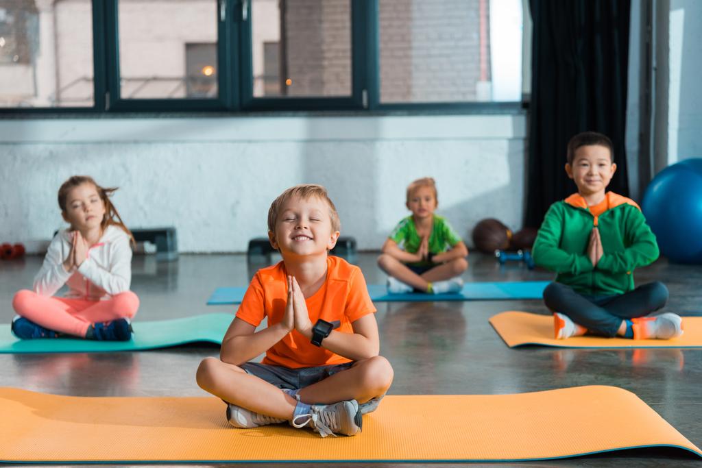 Selectieve focus van multi-etnische kinderen met gebalde handen en gekruiste benen zittend op fitnessmatten - Foto, afbeelding