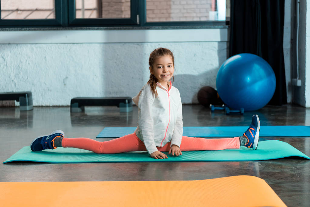 Foco seletivo da criança sorrindo e fazendo split no tapete de fitness no ginásio
 - Foto, Imagem