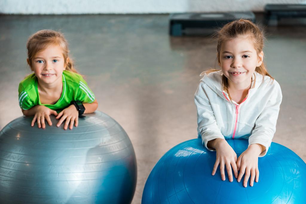 Селективный фокус детей, смотрящих в камеру, улыбающихся и опирающихся на фитнес-мячи в тренажерном зале
 - Фото, изображение