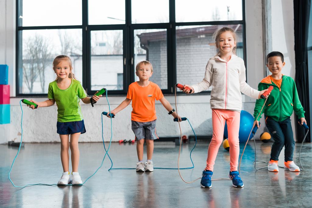 Vista frontal de niños multiétnicos haciendo ejercicio con cuerda de salto en el gimnasio
 - Foto, Imagen