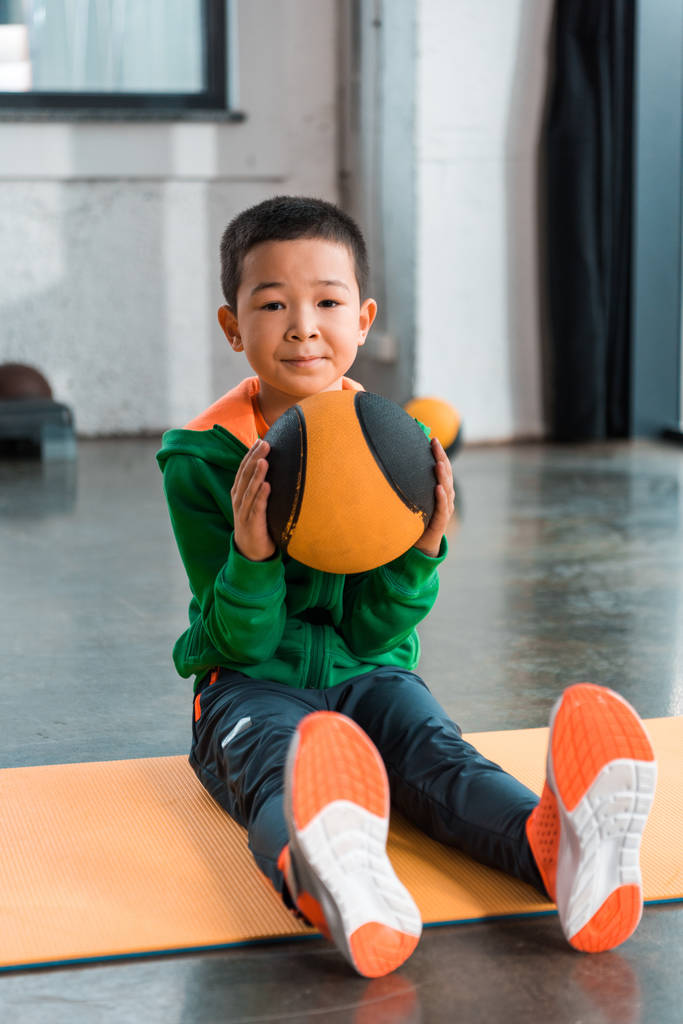 Selective focus of asian boy holding ball and sitting on fitness mat in gym - Photo, Image