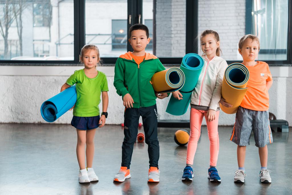 Widok z przodu Wielokulturowe dzieci trzymające maty fitness i patrzące w kamerę na siłowni - Zdjęcie, obraz
