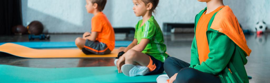 Enfoque selectivo de niños con piernas cruzadas sentados en colchonetas de fitness en el gimnasio, plano panorámico
 - Foto, imagen