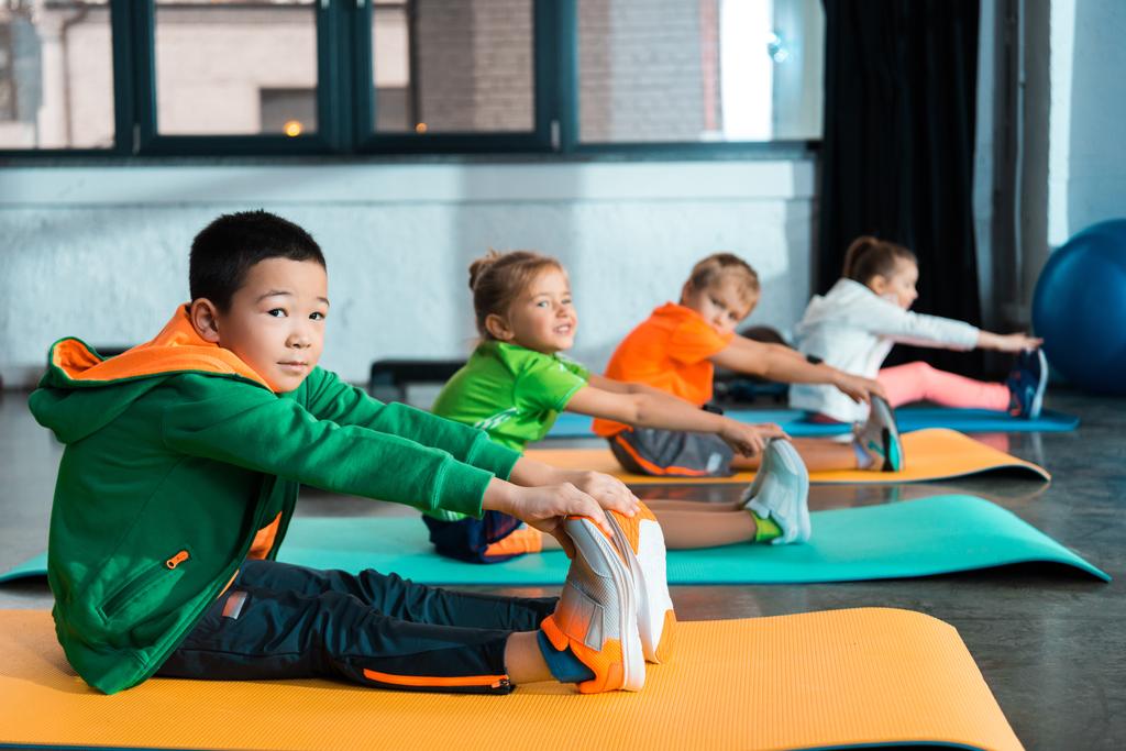 Foco seletivo de crianças multiétnicas que se alongam em tapetes de fitness no centro esportivo
 - Foto, Imagem