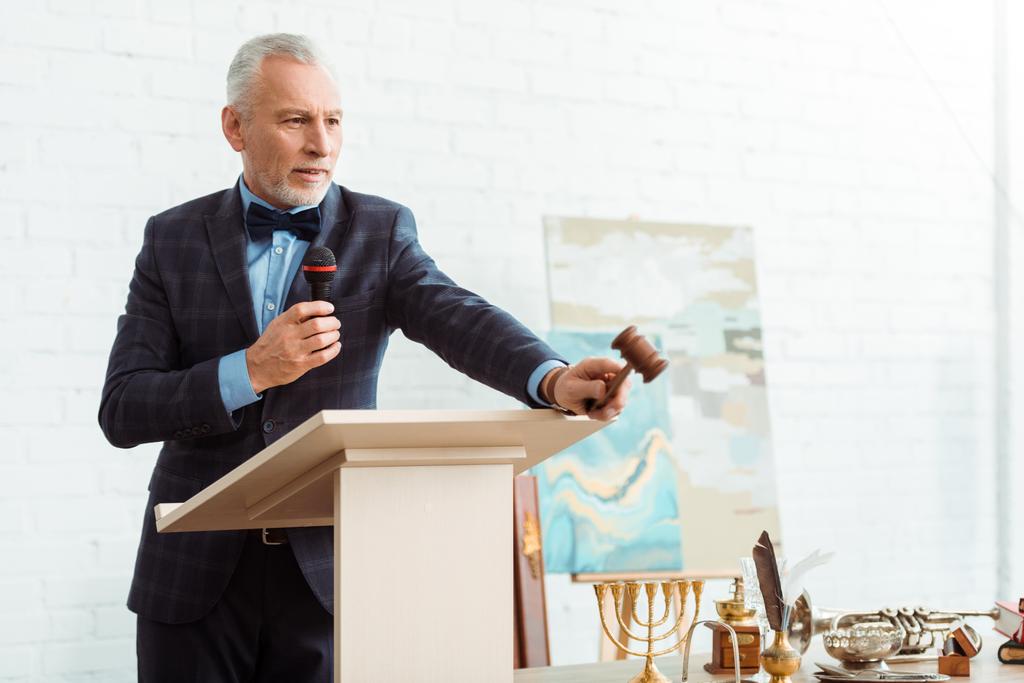 pohledný dražitel v obleku mluví s mikrofonem a drží kladívko během aukce - Fotografie, Obrázek