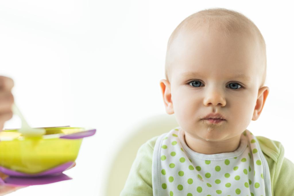 Selective focus of infant on feeding chair looking at camera near mother with bowl of puree isolated on white - Fotoğraf, Görsel