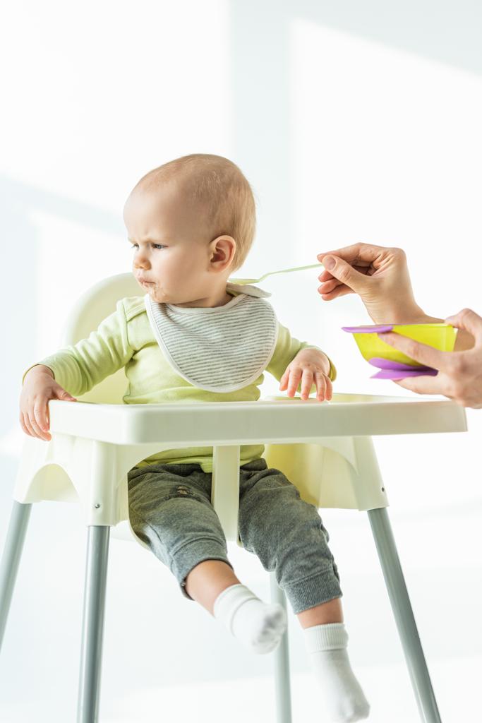 Baby op voederstoel weg te kijken in de buurt van moeder met kom van baby voeding en lepel op witte achtergrond - Foto, afbeelding