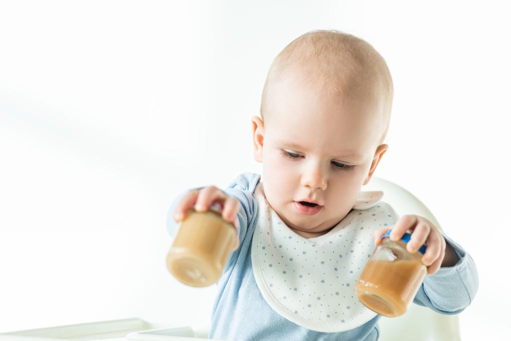 Enfoque selectivo del bebé en la silla de alimentación que sostiene frascos de nutrición del bebé de fruta sobre fondo blanco
 - Foto, imagen