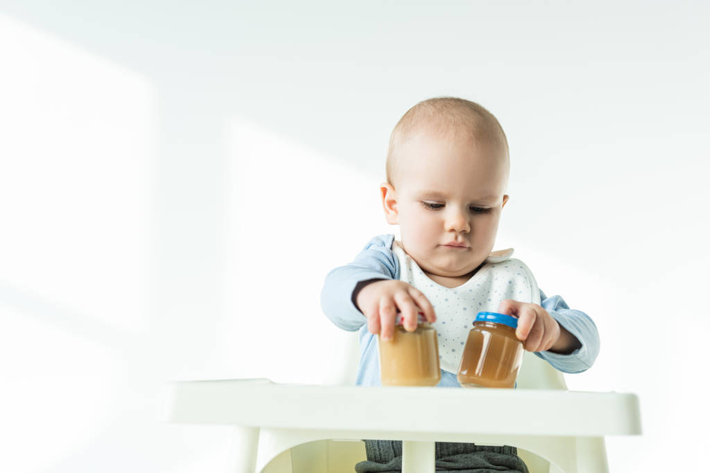 Cute baby holding jars of baby food on table of feeding chair on white background - Photo, Image
