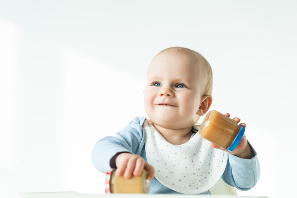 Selective focus of baby holding jars of fruit baby nutrition and smiling away while sitting on feeding chair on white background - Fotoğraf, Görsel