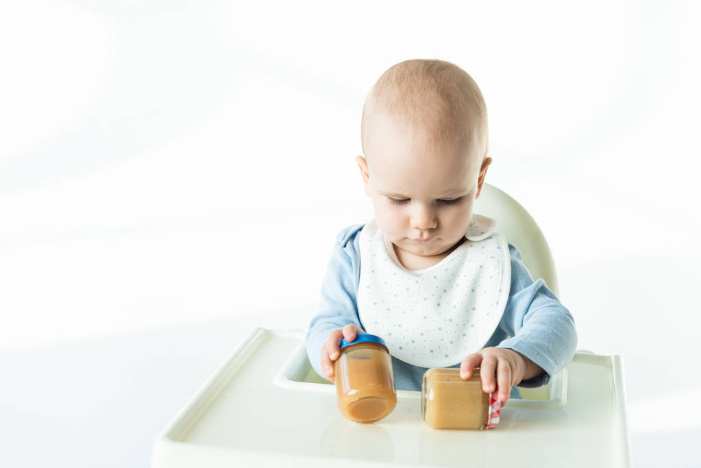 Adorable baby boy holding jars with baby food on table of feeding chair on white background - Fotoğraf, Görsel
