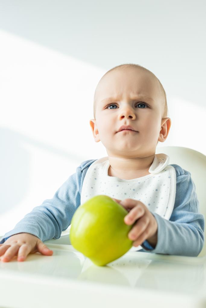 Focus selettivo del bambino pensoso che tiene la mela verde mentre siede sulla sedia di alimentazione su sfondo bianco
 - Foto, immagini