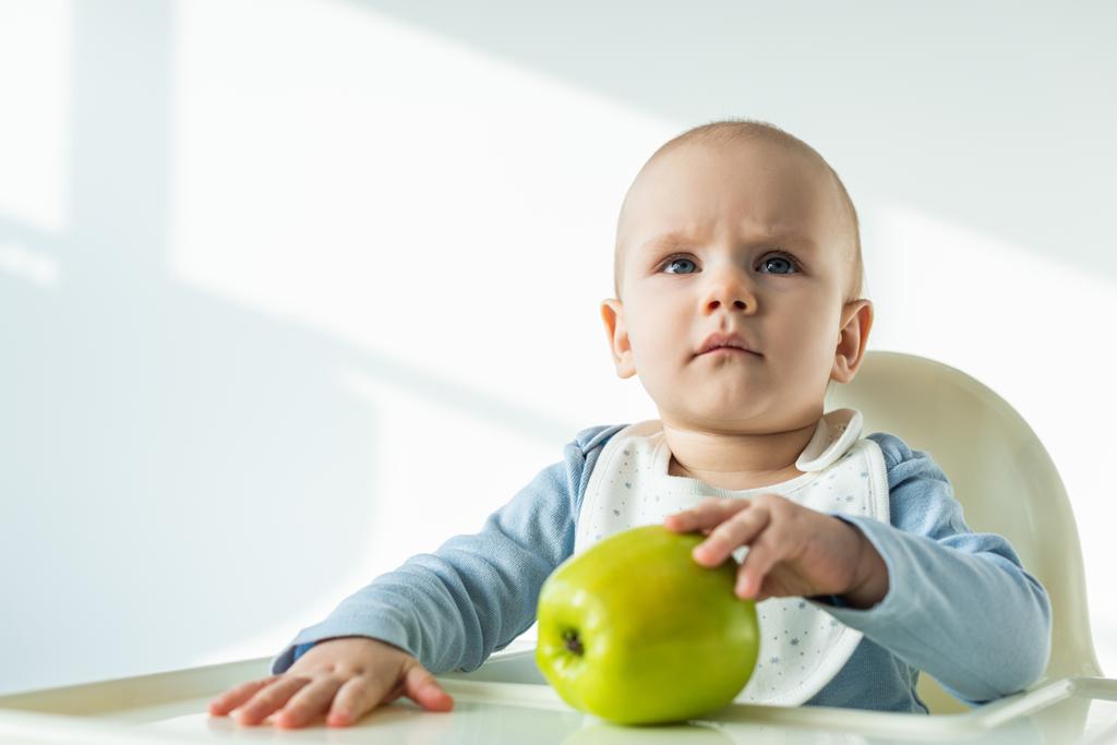 Cute baby boy touching green apple on table of feeding chair on white background - Photo, Image