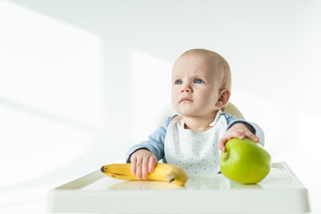 Adorable baby holding ripe banana and apple on table of feeding chair on white background - Photo, Image