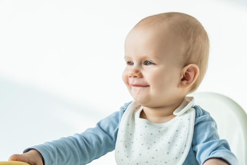 Glimlachende baby jongen met bevuilde mond zittend op voederstoel op witte achtergrond - Foto, afbeelding