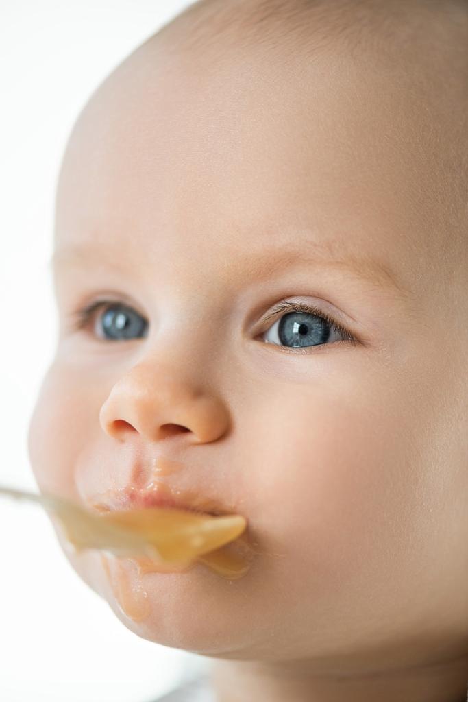 Enfoque selectivo de bebé lindo mirando hacia otro lado durante la alimentación con puré de frutas aislado en blanco
  - Foto, Imagen