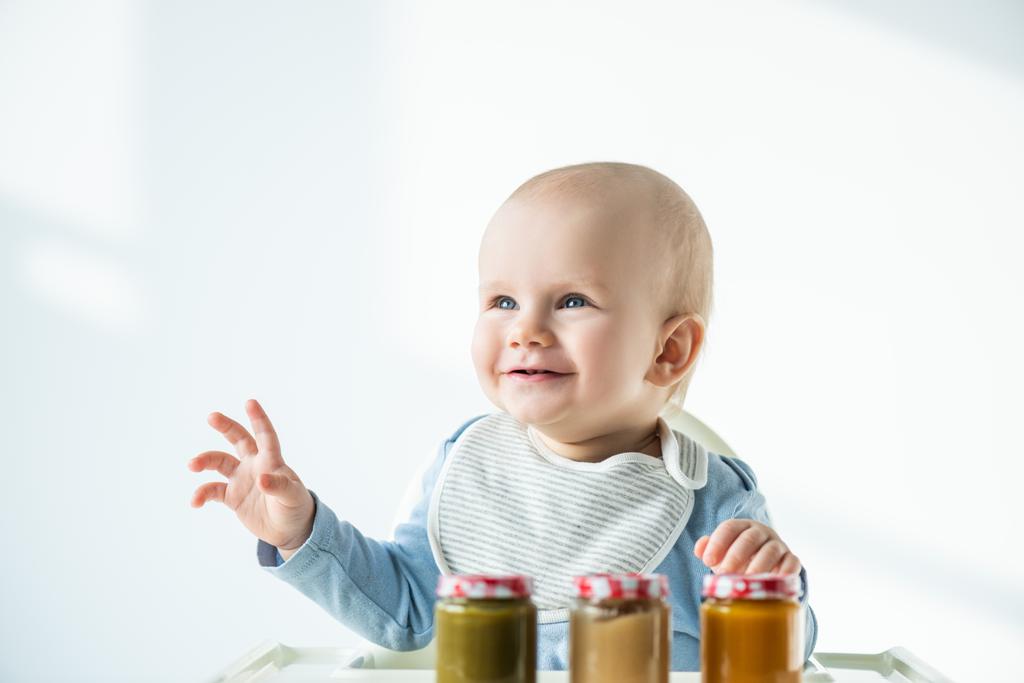 Selective focus of baby boy smiling away near jars of baby nutrition on table of feeding chair on white background - 写真・画像