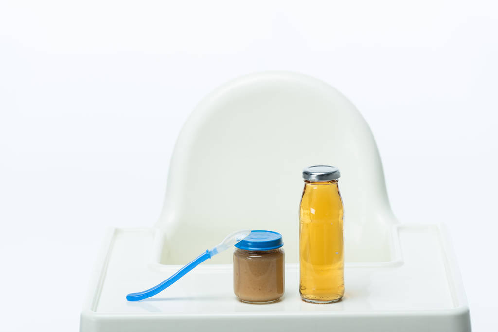 Tarro de comida para bebés con cuchara y biberón de jugo de manzana en la mesa de la silla de alimentación aislado en blanco
 - Foto, imagen