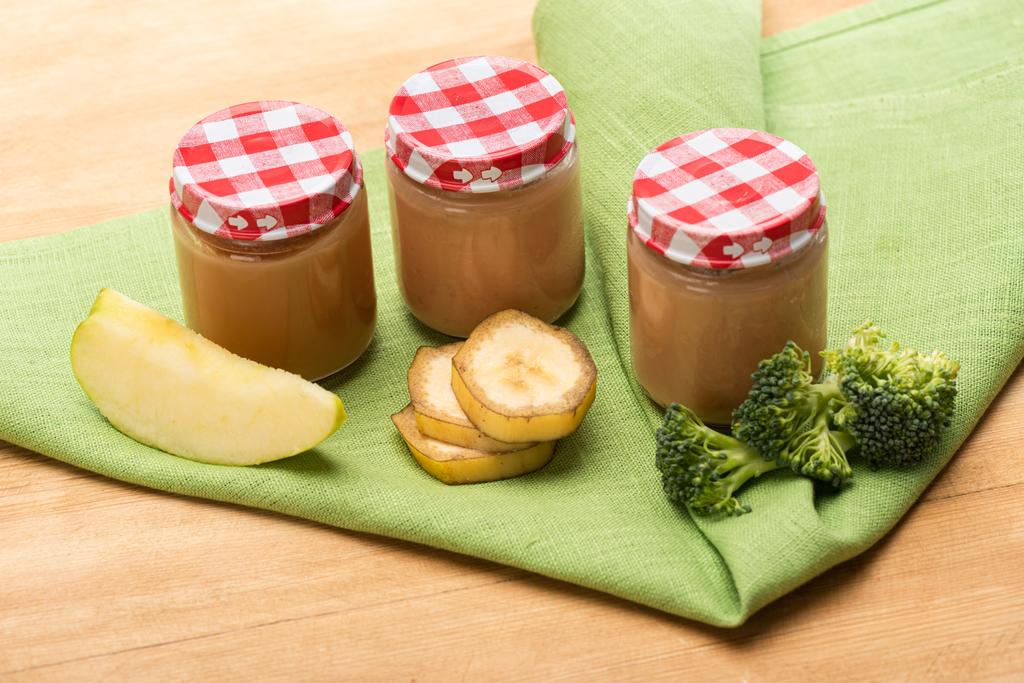 Potten bereide babyvoeding met stukjes appel, broccoli en bananenschijfjes op servet op houten ondergrond - Foto, afbeelding