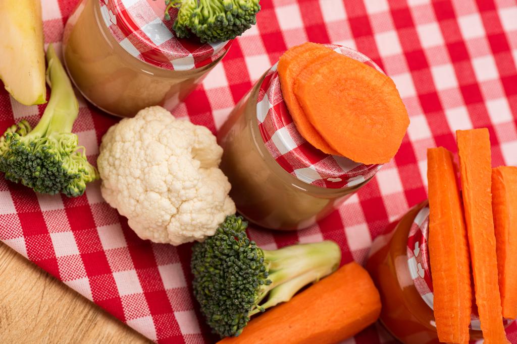 High angle view of jars of vegetable baby food with fresh vegetables and apple on tablecloth on wooden surface - 写真・画像