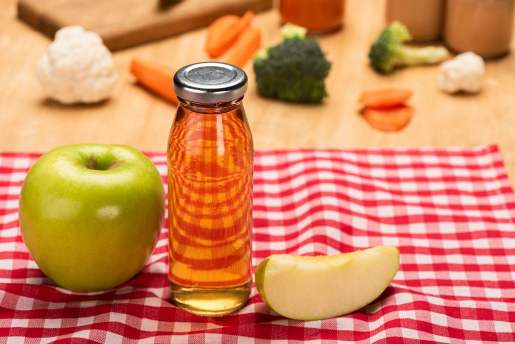 Enfoque selectivo del biberón de jugo de manzana y alimentos para bebés en frascos con ingredientes en mantel sobre fondo de madera
 - Foto, Imagen