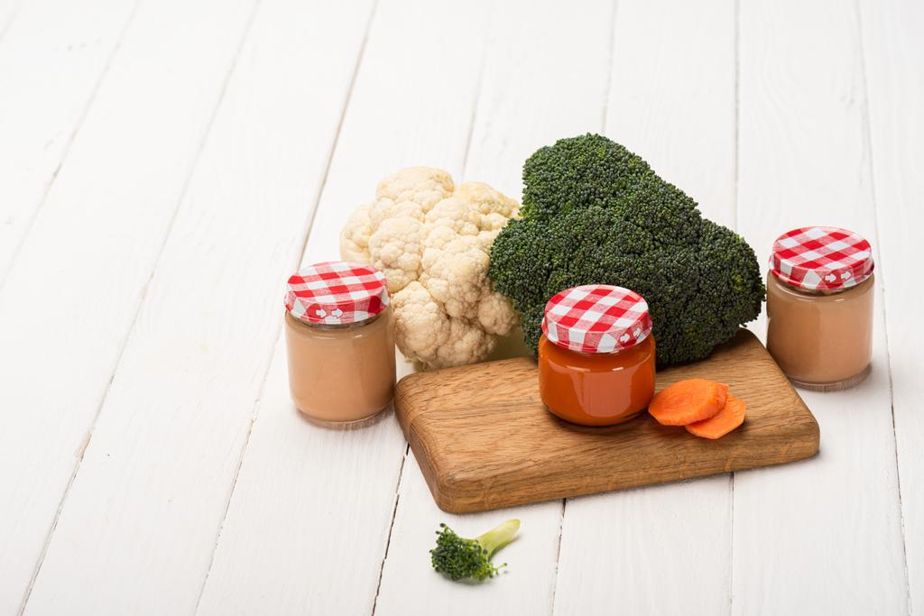 Pots d'aliments pour bébés avec des tranches de brocoli, de chou-fleur et de carotte sur la planche à découper sur une surface en bois blanc
 - Photo, image