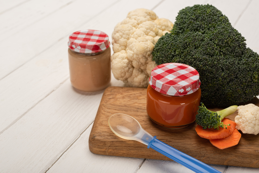 Vista de cerca de la cuchara con frascos de nutrición para bebés y verduras frescas en la tabla de cortar en la superficie de madera blanca
 - Foto, imagen